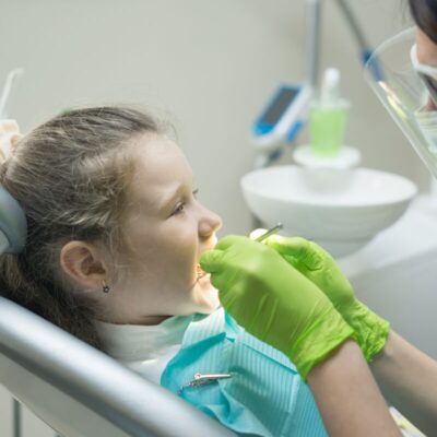 Dentystka pracuje z dzieckiem na fotelu dentystycznym, przypominając o różnicach między znieczuleniem ogólnym a podtlenkiem azotu stosowanym w stomatologii.