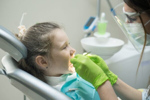Dentystka pracuje z dzieckiem na fotelu dentystycznym, przypominając o różnicach między znieczuleniem ogólnym a podtlenkiem azotu stosowanym w stomatologii.