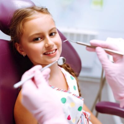 Dziewczynka siedząca na fotelu dentystycznym podczas badania, zadając sobie pytanie, czy wizyta adaptacyjna jest konieczna.