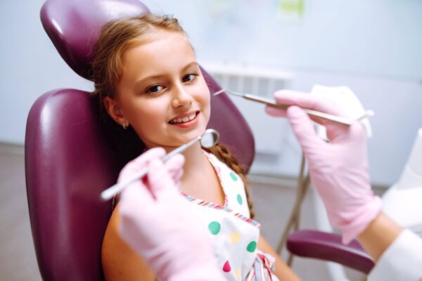 Dziewczynka siedząca na fotelu dentystycznym podczas badania, zadając sobie pytanie, czy wizyta adaptacyjna jest konieczna.